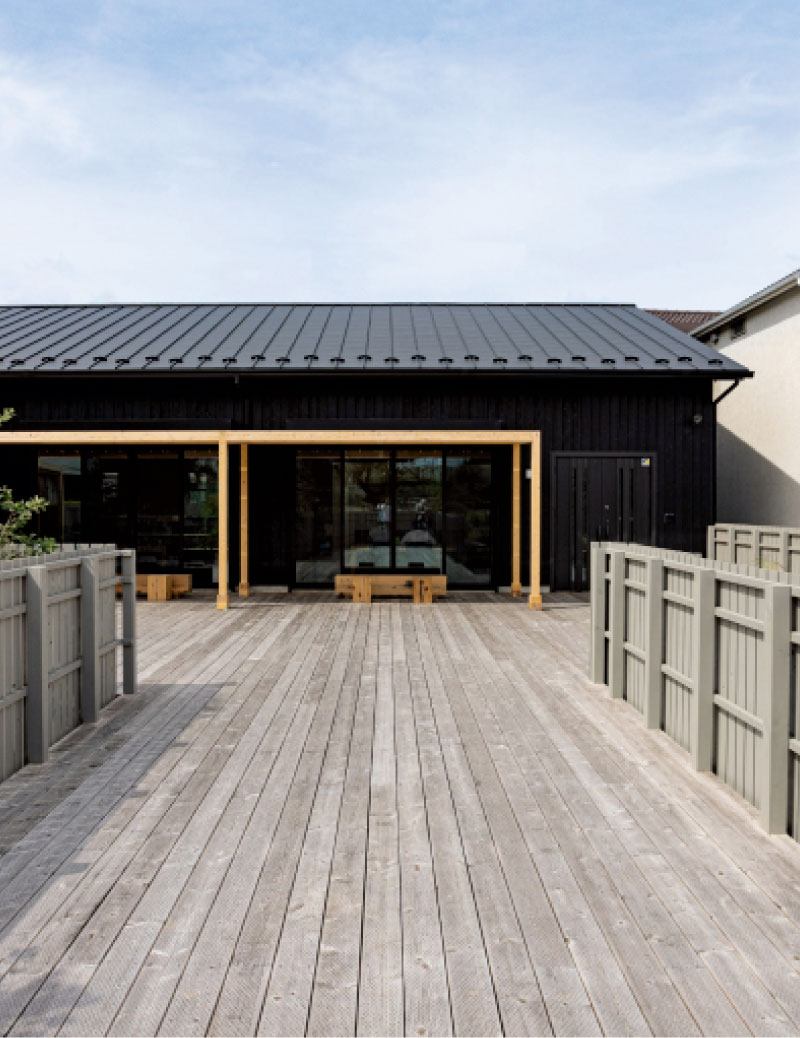 外観。黒い外壁と屋根の建物の前に広い木製デッキが設けられている。